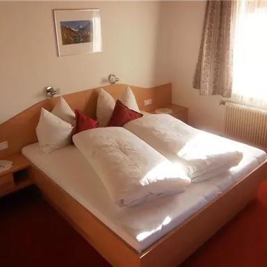 Ein gemütliches Schlafzimmer mit einem großen Doppelbett und zwei Kissen. Das Zimmer ist hell und freundlich, mit einem Fenster, durch das Sonnenlicht hereinströmt.