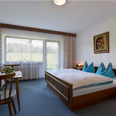 Ein gemütliches Schlafzimmer mit zwei Betten und blauen Kissen. Große Fenster bieten einen Blick auf die grüne Landschaft.