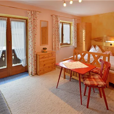 Ein gemütliches Zimmer mit Holzmöbeln und einer kleinen Sitzecke. Große Fenster lassen viel Licht hereinscheinen.