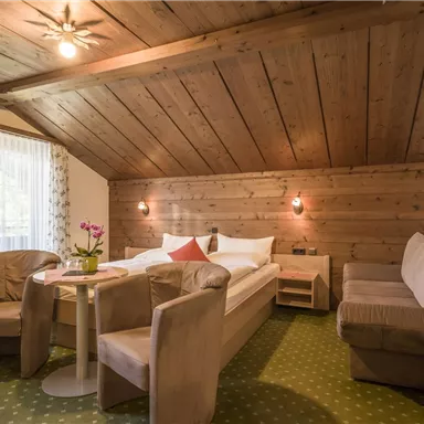 Ein gemütliches Zimmer mit Holzverkleidung und einem Dachschrägen. Es gibt eine Sitzecke mit einem Tisch sowie ein Doppelbett.