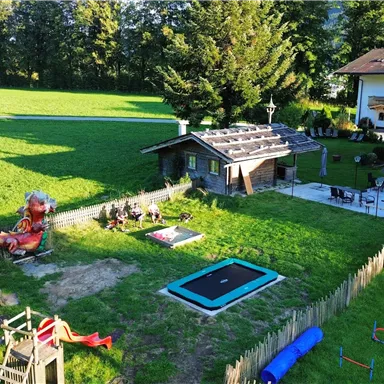 Ein großer Garten mit einem Spielplatz und einem Trampolin. Im Hintergrund steht ein Haus, umgeben von Bäumen und einer grünen Wiese.