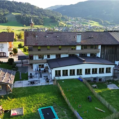 Ein malerisches Gebäude inmitten von grüner Landschaft. Im Vordergrund befindet sich ein Garten mit Spielmöglichkeiten.