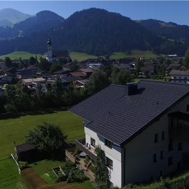 A picturesque alpine landscape with a village and tall mountains in the background. In the foreground, there is a modern house with a green garden.
