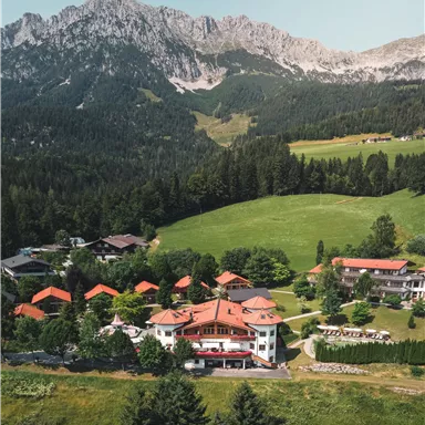 Ein malerisches Hotel inmitten von grünen Wiesen und Wäldern, umgeben von majestätischen Bergen. Die roten Dächer der Gebäude strahlen eine einladende Atmosphäre aus.