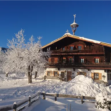 Ein traditionelles Holzhaus in einer verschneiten Landschaft. Umgeben von schneebedeckten Bäumen und klarem, blauem Himmel.