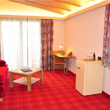 A cozy room with a red carpet and curtains. There is a small table, a chair, and a refrigerator.