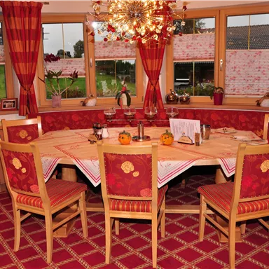 A cozy dining area with a round wooden table and striped chairs. Large windows offer a view of the greenery and are decorated with red curtains.