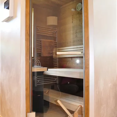 A modern sauna with a glass door and wooden shelves. In the foreground, there is a wooden container for water.
