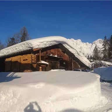 Eine verschneite Landschaft mit einem traditionellen Holzhaus und Bergen im Hintergrund. Der Himmel ist klar und blau, was eine ruhige Winteratmosphäre schafft.