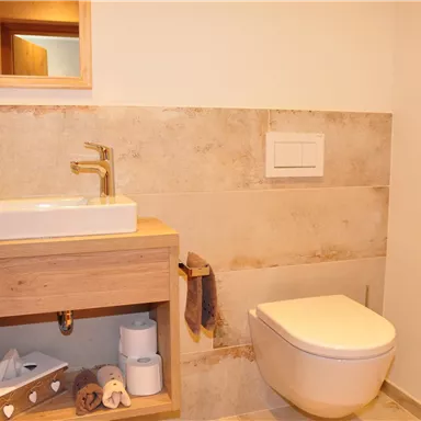 A modern bathroom with a sink on a wooden shelf and a toilet. The walls are in neutral colors and there is a small wooden decor.