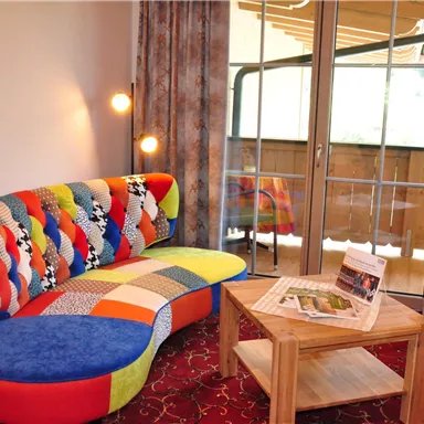 A cozy living area with a colorful upholstered sofa and a wooden table. Large windows provide a view of the balcony.