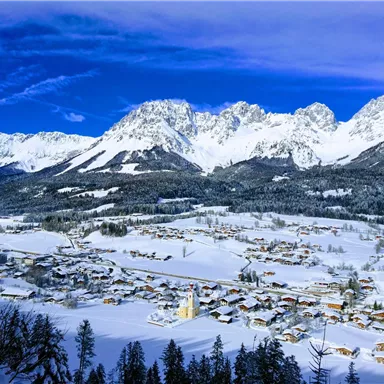 A picturesque winter landscape with snow-covered mountains and a quiet village. The sky is bright blue and the scene is peaceful.