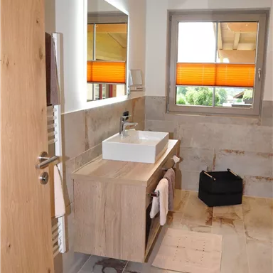 A modern bathroom with a white sink and wooden shelf. Large windows provide plenty of natural light, and the orange blinds add a touch of accent.