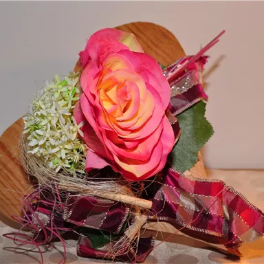 A beautiful bouquet of a pink rose and white flowers, decorated with a colorful ribbon. The flowers are arranged on a wooden heart, creating an appealing presentation.