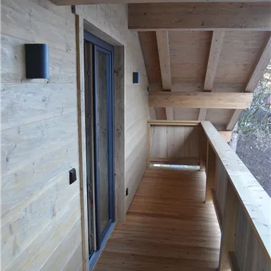 Ein schmaler Balkon mit Holzverkleidung und einem großen Fenster. Die Umgebung ist ruhig und gemütlich, ideal zum Entspannen.