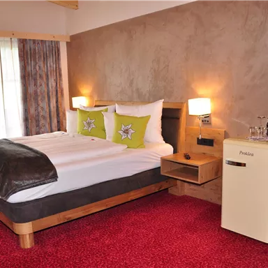 A cozy bedroom with a large bed, colorful pillows, and a small refrigerator. The room has a red carpet and large windows that let in plenty of light.