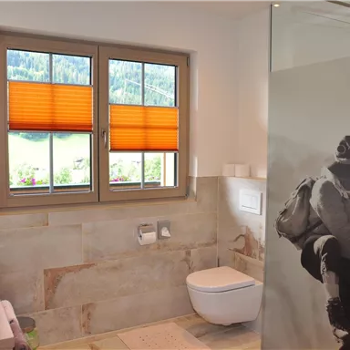 A modern bathroom with two sinks and a toilet area. Large windows with orange blinds provide daylight and a view of nature.