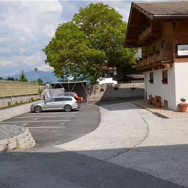 Ein Parkplatz neben einem Holzgebäude mit einem großen Baum im Hintergrund. Der Himmel ist teils bewölkt.