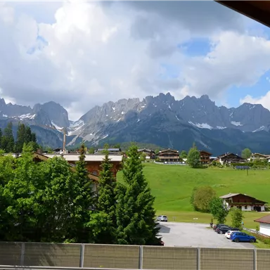 Eine malerische Berglandschaft mit hohen Bergen und grünem Gras. Im Vordergrund sind Bäume und einige Wohnhäuser zu sehen.