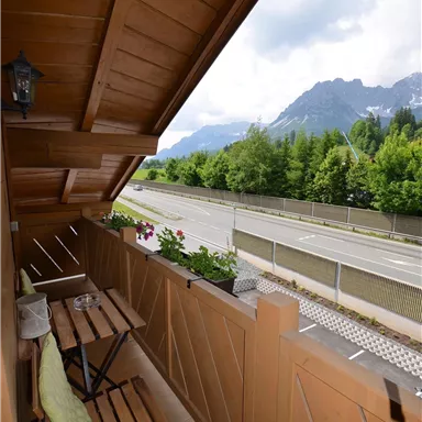 Ein Balkon mit Holzmöbeln und Blumenkästen. Im Hintergrund sieht man eine Straße und beeindruckende Berge.