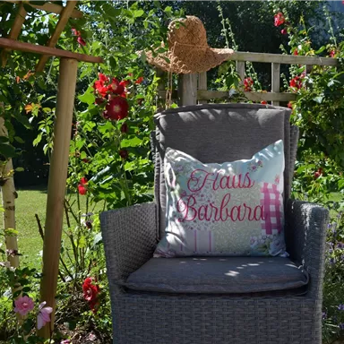 Ein bequemer Stuhl im Garten, umgeben von bunten Blumen. Auf dem Stuhl liegt ein Kissen mit der Aufschrift "Haus Barbord".