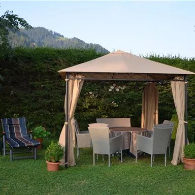 Ein gemütlicher Garten mit einem Pavillon und einem Esstisch. Im Hintergrund sind Blumen und ein schattiger Liegestuhl zu sehen.