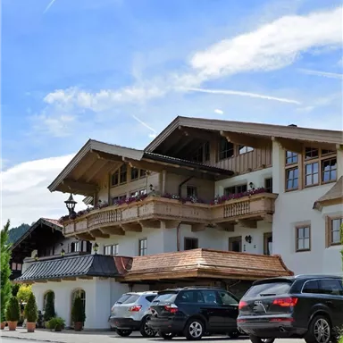 Ein schönes alpines Gebäude mit traditioneller Architektur und einem großzügigen Balkon. Vor dem Gebäude stehen mehrere Autos geparkt.