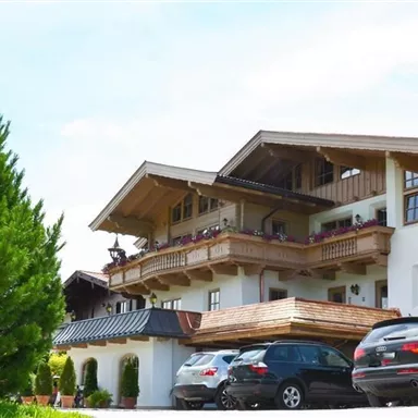 Ein charmantes alpenländisches Gebäude mit Holzverzierung und Balkonen. Vor dem Haus stehen mehrere Autos und es gibt viel grünes Umfeld.