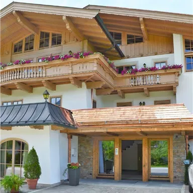 Ein gemütliches Holzhaus mit Balkonen und bunten Blumen. Die Vorderansicht zeigt den Eingangsbereich und eine gepflasterte Auffahrt.