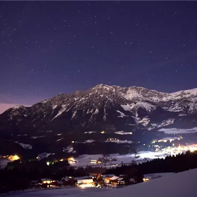 A winter landscape at night with snow-covered mountains and a clear starry sky. Small lights from villages gently glow in the snow.