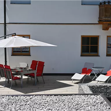 A cozy outdoor area with a table and red chairs under a sun umbrella. There are also two loungers and a modern setting made of stones.