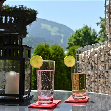 A table outside with two glasses of drinks and a lantern. In the background, green mountains and a clear sky can be seen.