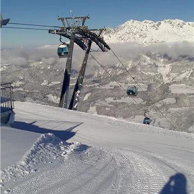 A cable car with blue gondolas in a snow-covered landscape. In the background, there are high mountains and a clear sky.