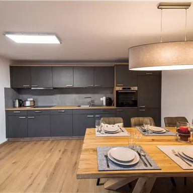 Eine moderne Küche mit einem großen Esstisch aus Holz. Die Wände sind in einem neutralen Farbton gehalten, und die Ausstattung ist elegant und funktional.