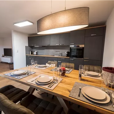 Ein modernes Esszimmer mit einem großen Holztisch und stilvollen Stühlen. Die offene Küche im Hintergrund ist funktional und gut ausgestattet.