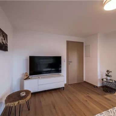 Ein modernes Wohnzimmer mit einem Fernseher und einem Holztisch. Die Wände sind hell, und der Boden ist aus Holz.