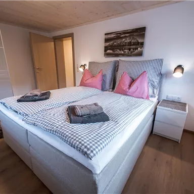 Ein stilvolles Schlafzimmer mit einem großen Bett und rosa Kissen. Der Raum hat einen modernen Look mit Holzfußboden und schlichten Möbeln.