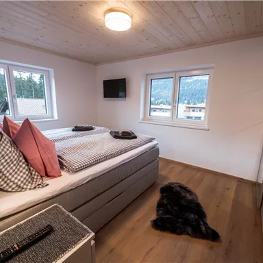 Ein helles Schlafzimmer mit einem großen Bett und mehreren Kissen. Es gibt zwei Fenster, einen Fernseher an der Wand und einen weichen Teppich auf dem Boden.