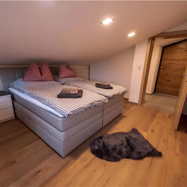 Ein gemütliches Schlafzimmer mit zwei Betten und Kissen. Der Raum hat einen Holzfußboden und eine offene Tür, die zu einem weiteren Bereich führt.