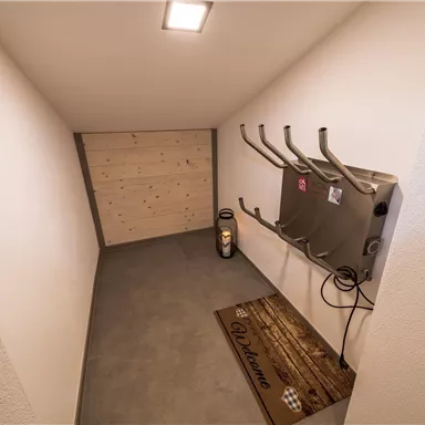 Ein kleiner, heller Raum mit einem grauen Fußboden und einer Holzwand. An der Wand hängen mehrere Halterungen und es steht ein Elektrowärmer auf dem Boden.