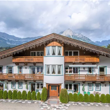 A traditional Alpine house with decorative balconies and plenty of greenery surrounding it. In the background, majestic mountains and a cloudy sky can be seen.