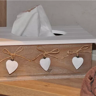A decorative tissue box made of wood, adorned with hearts. Next to it lies a towel.