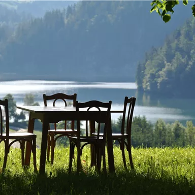 A table with chairs is set on a meadow overlooking a lake. In the background, there are trees and gentle hills.