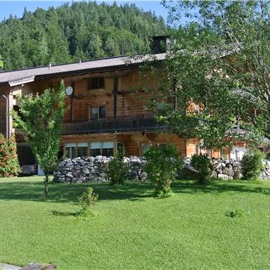 A rustic wooden house surrounded by a green garden. In the background, gentle hills with trees can be seen.