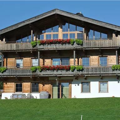 A traditional wooden house with several balconies and colorful flowers. It is located in a green environment under a blue sky.