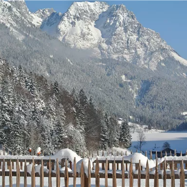A winter mountain view with snow-covered peaks and a frozen lake. In the foreground, there is a wooden fence that frames the beautiful landscape.