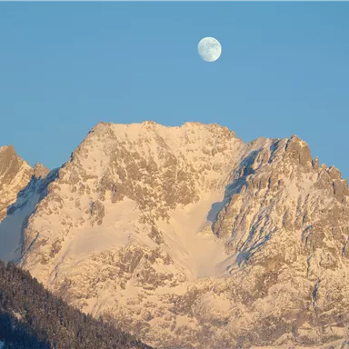 A majestic mountain with snow-covered peaks under a clear blue sky. The moon shines over the mountains.