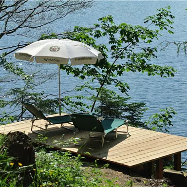 A cozy terrace by the water with loungers and a sun umbrella. Surrounded by green trees and calm water.