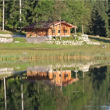 A beautiful wooden house by the shore of a tranquil lake, surrounded by trees. The image shows the reflection of the house in the water.