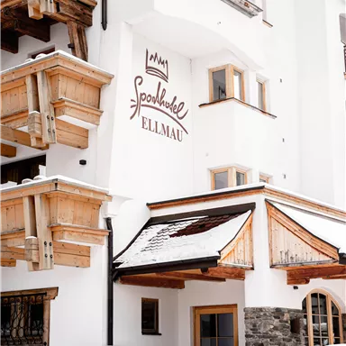 A charming hotel with rustic wooden balconies and an inviting facade. The surroundings are wintry with a blanket of snow.